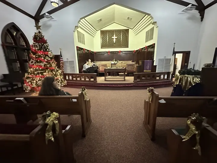 The sanctuary at Christmas with a lit tree, poinsettias along the aisle, and garland on the pews.