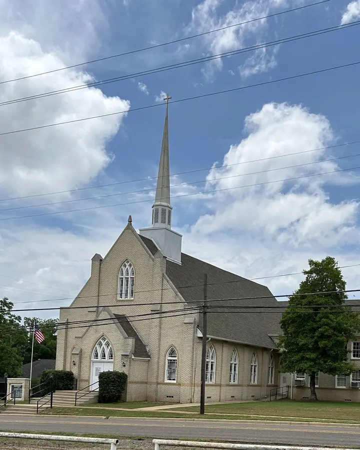The brick sanctuary and tall white steeple of Hughes Springs Methodist Church beneath a blue Texas sky.