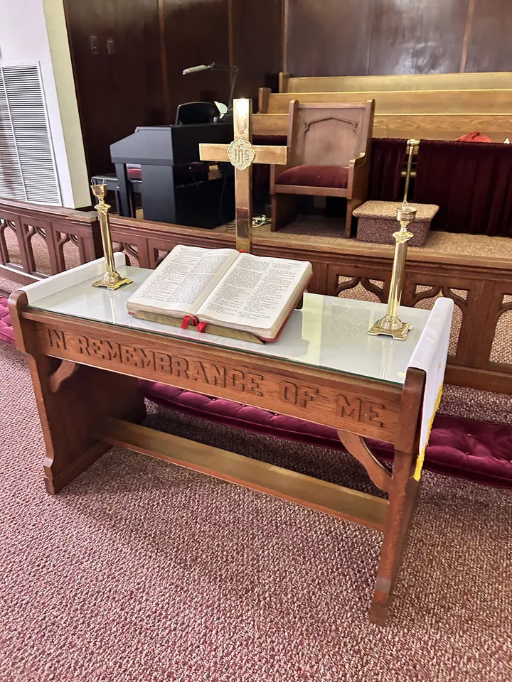 A carved wooden communion table inscribed 'In Remembrance of Me,' holding an open Bible between two brass candlesticks and a brass cross.
