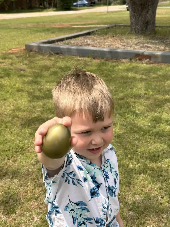 A young boy grinning as he holds up a gold Easter egg on the church lawn.