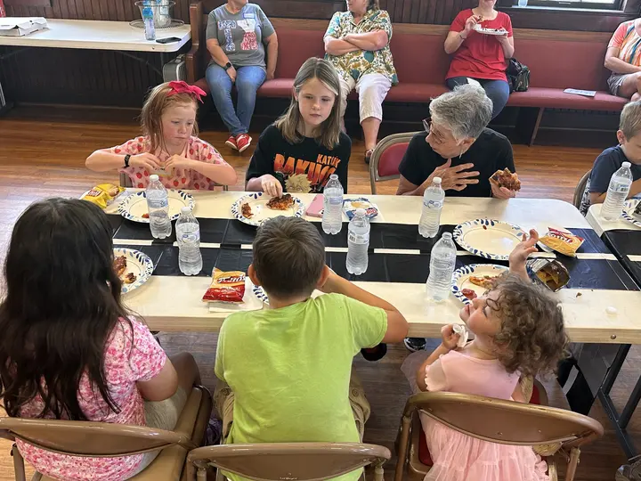 Children and adults seated together at long tables in the fellowship hall sharing a meal.