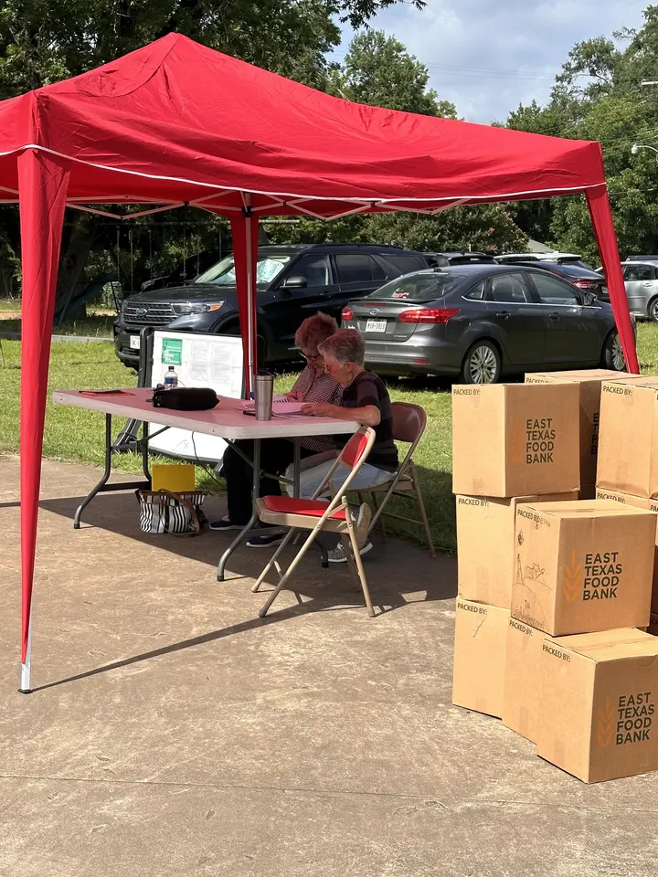 A volunteer working under a red canopy beside stacked East Texas Food Bank boxes on distribution day.