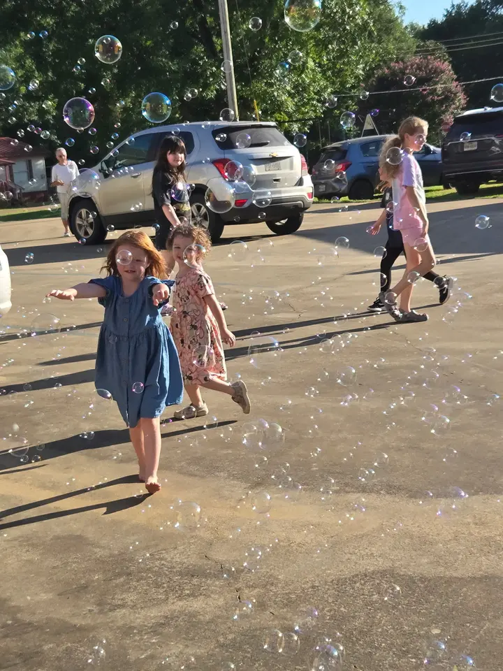 Children laughing as they chase soap bubbles across the church parking lot on a summer evening.