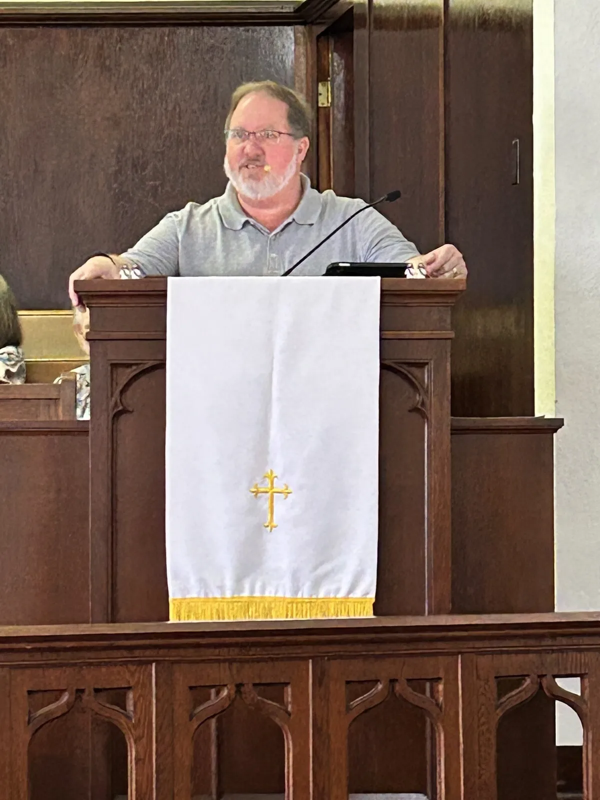Pastor Chad Mitchell preaching from the wooden pulpit beside a white parament with a gold cross.