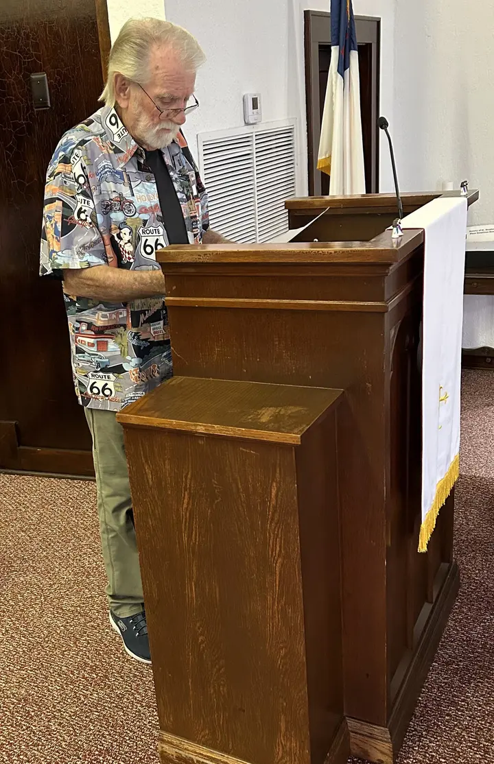A lay reader leading worship from the carved wooden pulpit, with a Texas flag behind.
